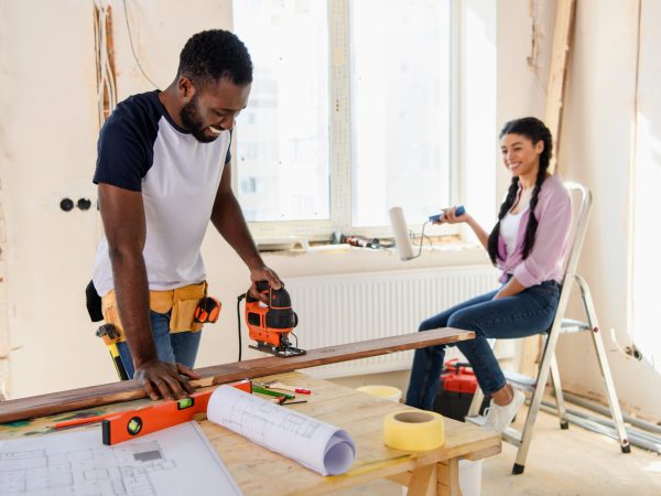smiling african american man working with jigsaw while his girlfriend sitting near during renovation